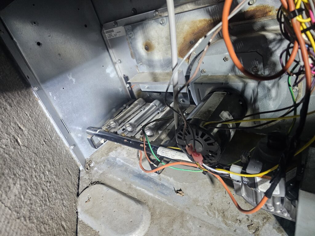 Interior view of a gas furnace showing burner assembly, inducer draft motor, heat exchanger tubes, and wiring with visible rust and heat discoloration on the metal cabinet