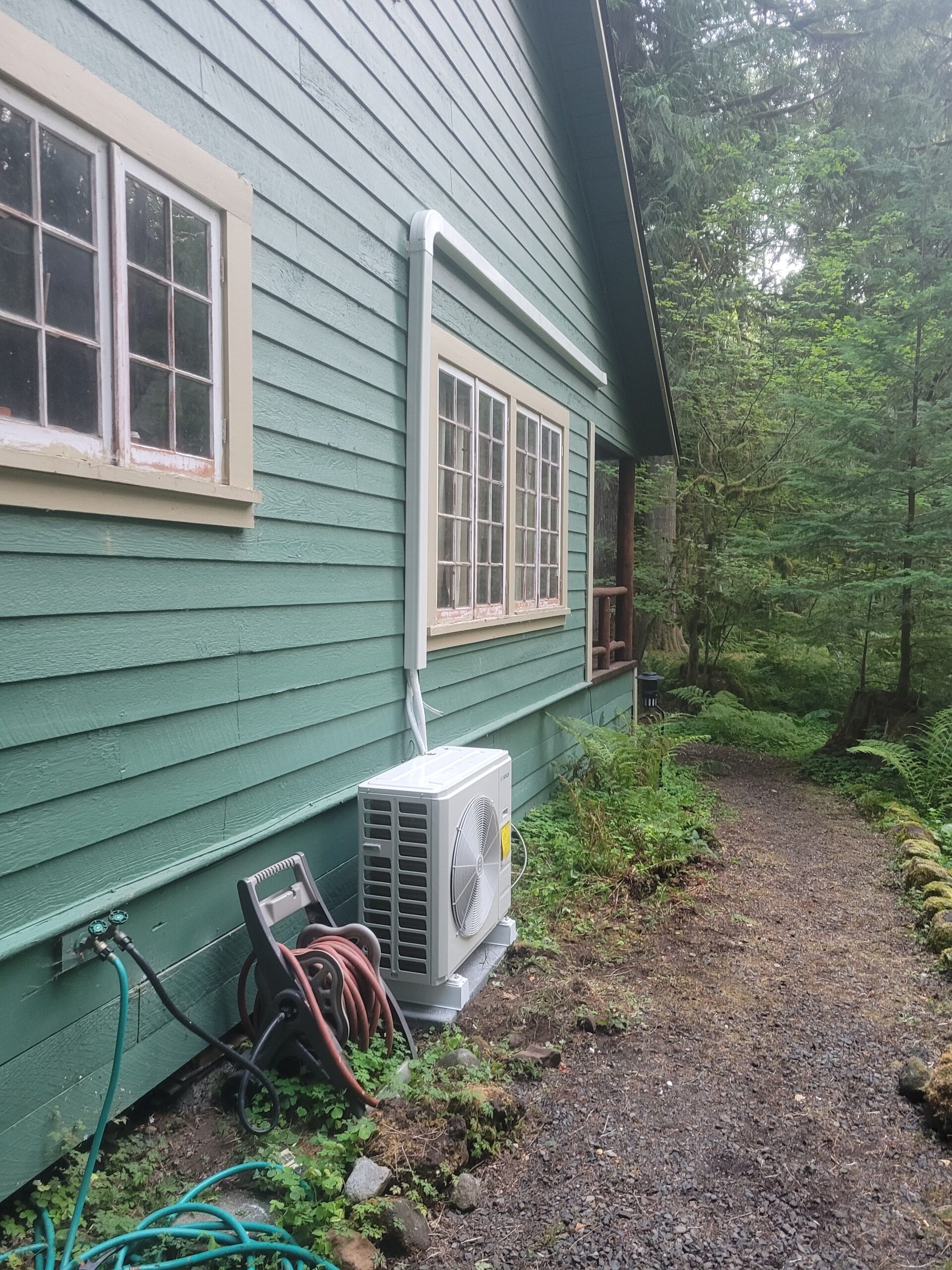 Ductless mini-split heat pump outdoor unit mounted on a green wood-sided home in a Pacific Northwest setting