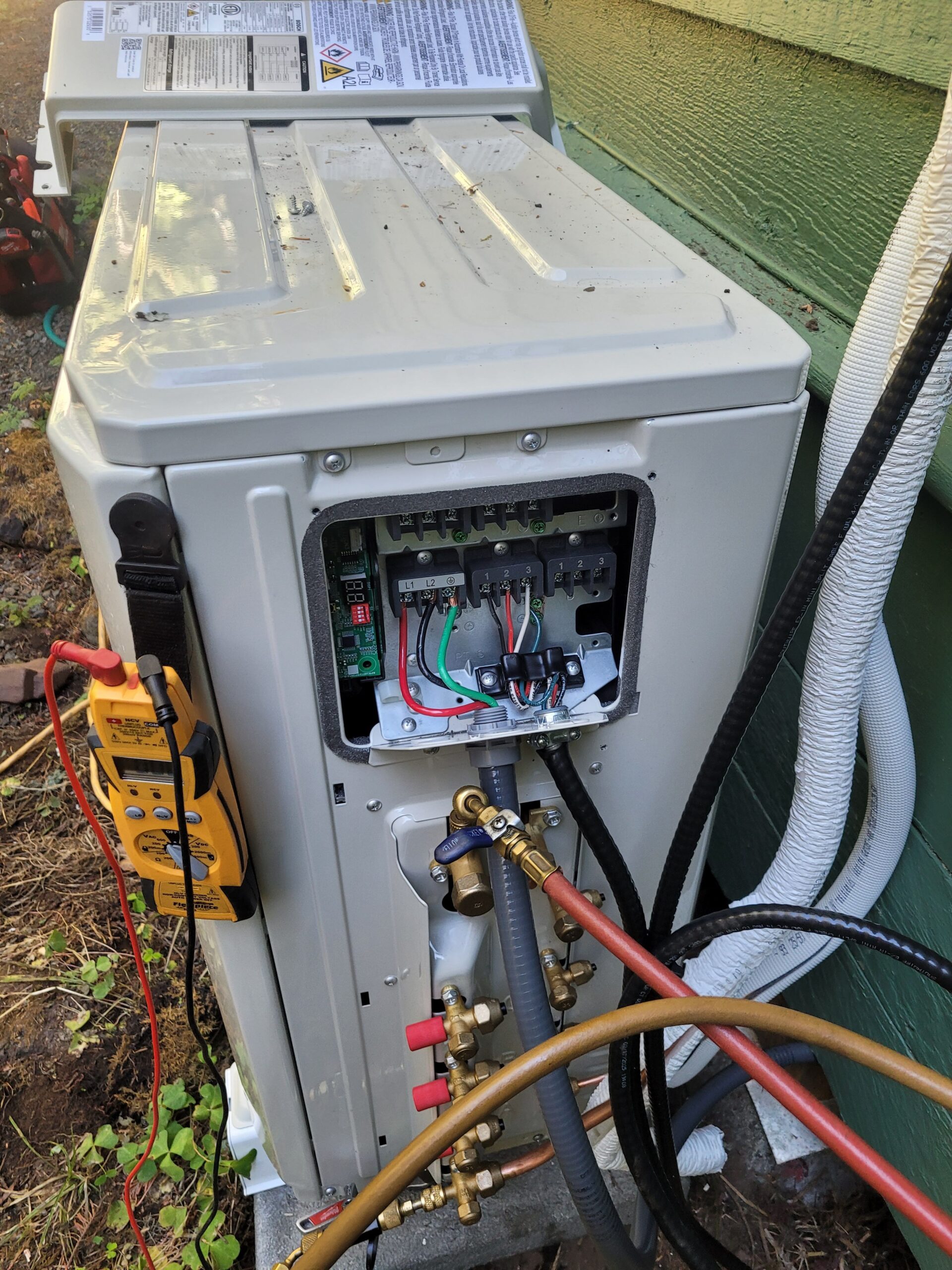 Mini-split outdoor unit with electrical panel open, manifold gauges on the service valves and a multimeter connected for testing