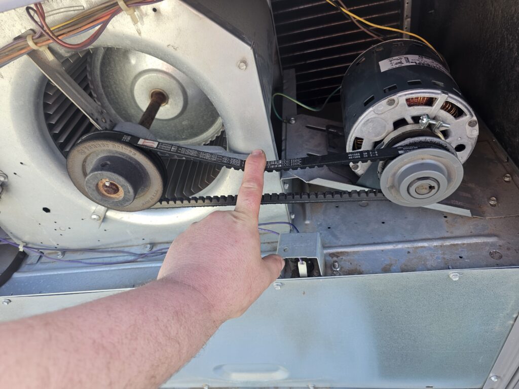 Technician's finger pressing a cogged drive belt between a blower pulley and electric motor inside an HVAC unit