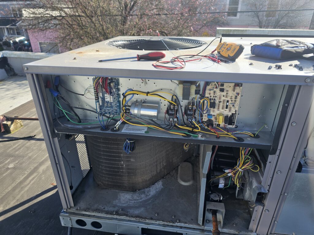 Open rooftop packaged HVAC unit showing control board, capacitor, contactors and wiring, with a multimeter on top