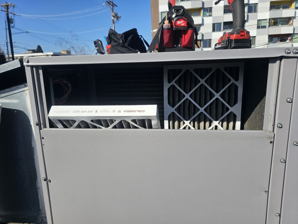 Rooftop HVAC unit with access panel open showing pleated air filters, a new filter going in, and tools on top