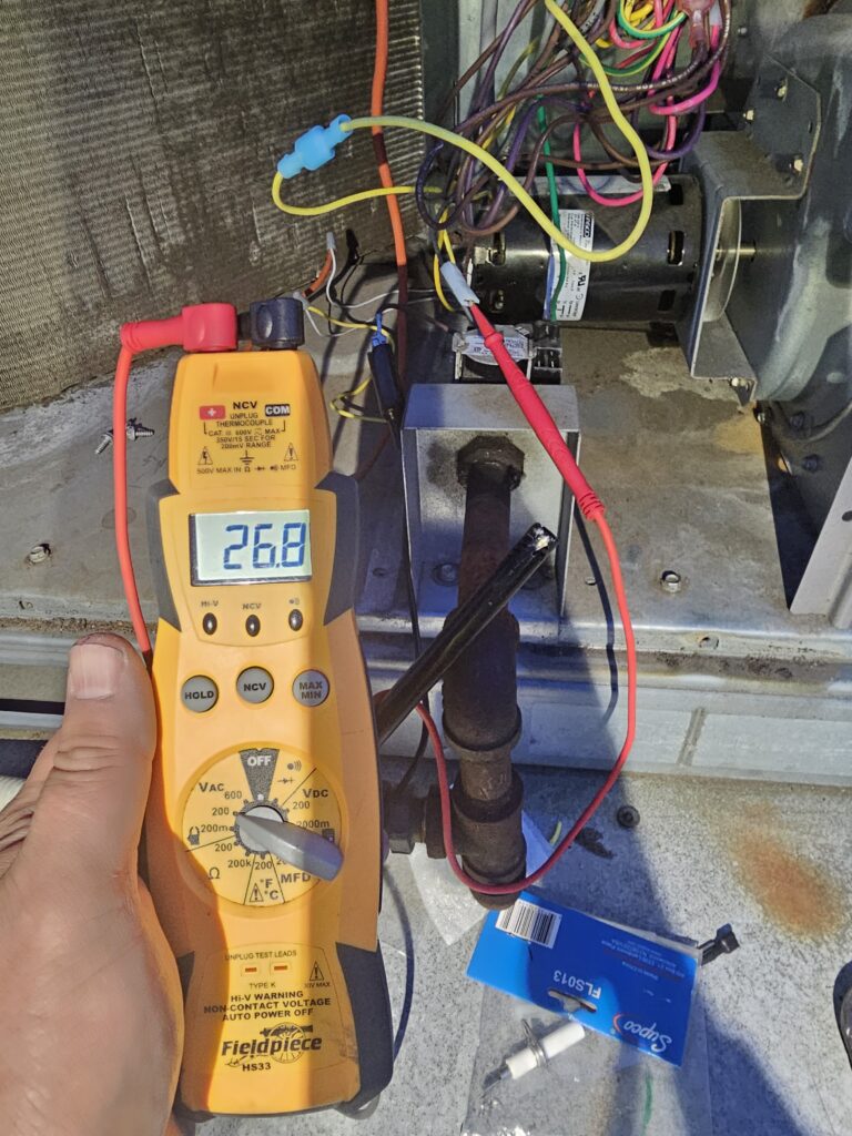 Fieldpiece multimeter reading 26.8 with test leads on a gas heating unit wire, beside a new Supco flame sensor package