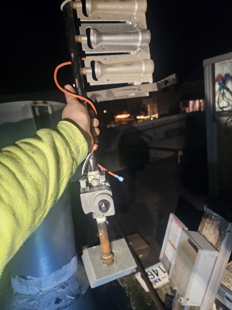 Technician in a hi-vis sleeve holds a removed gas burner assembly with gas valve and wiring on a rooftop at night