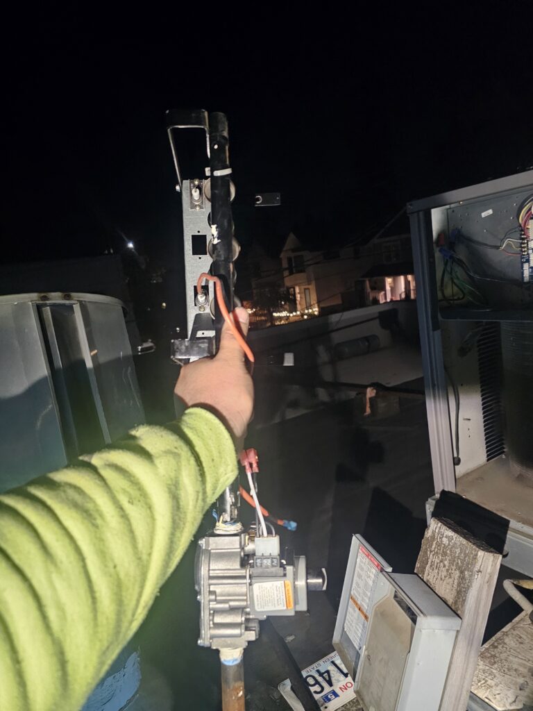 Technician holding a gas burner manifold and valve pulled from a rooftop HVAC unit at night, open unit panel at right