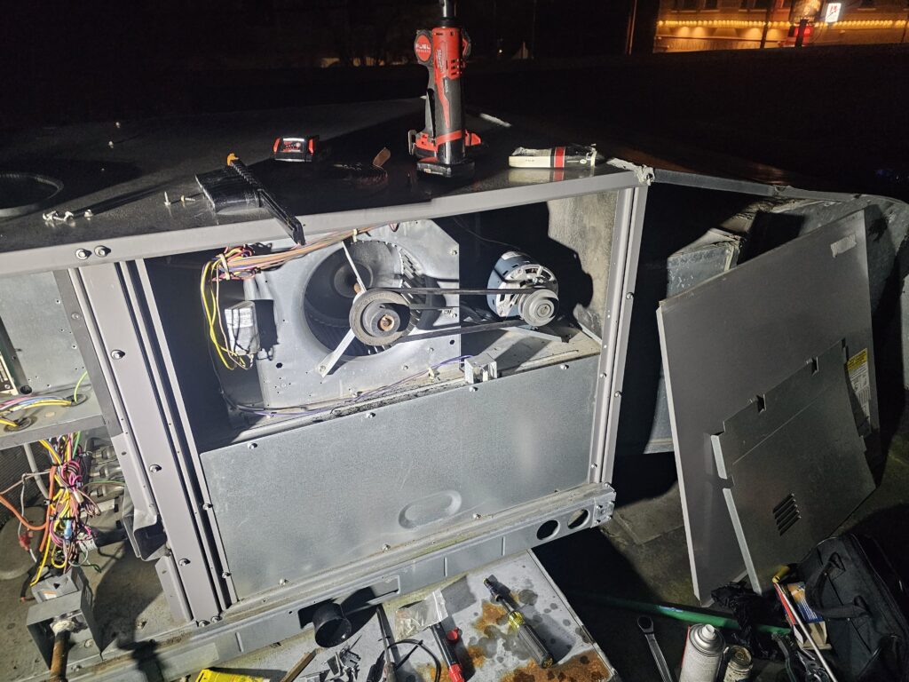Rooftop HVAC unit at night with access panel off, showing belt-driven blower, motor pulley, and wiring, with tools nearby