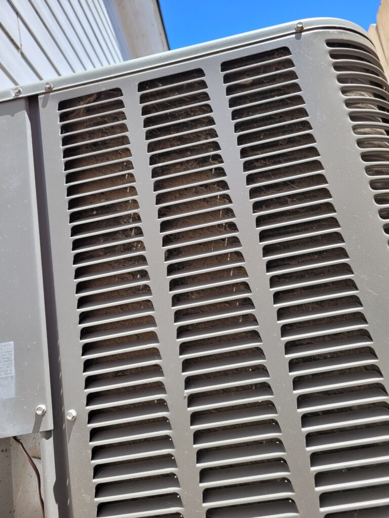 Gray outdoor AC condenser unit with louvered side panels and a coil behind them caked with dust, lint, and debris