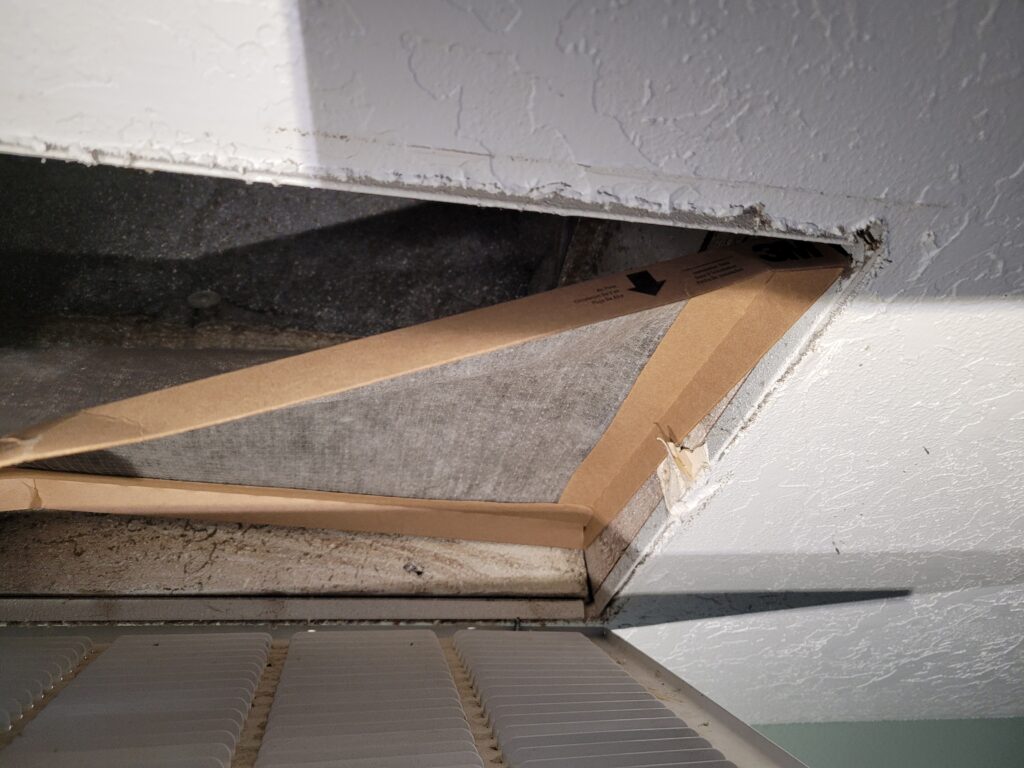 Open ceiling return air grille revealing a dust-clogged, collapsed air filter with a cardboard frame inside a dirty duct opening