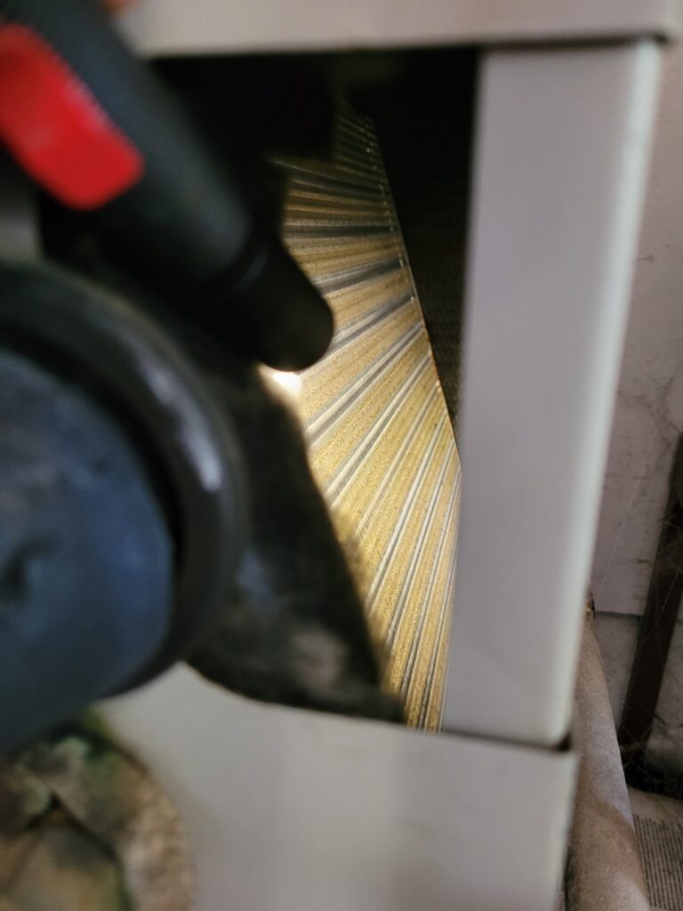Spray nozzle with red trigger aimed at aluminum condenser coil fins through an opening in a beige outdoor AC cabinet