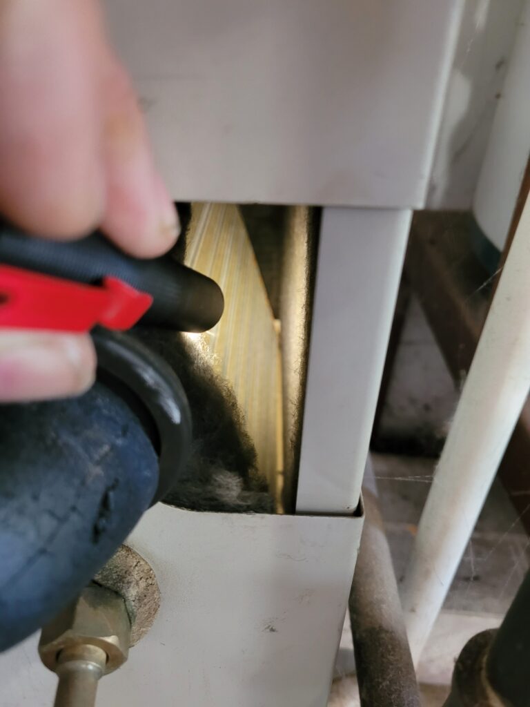 Hand holding a vacuum hose to remove thick dust and lint from coil fins inside a white HVAC cabinet opening