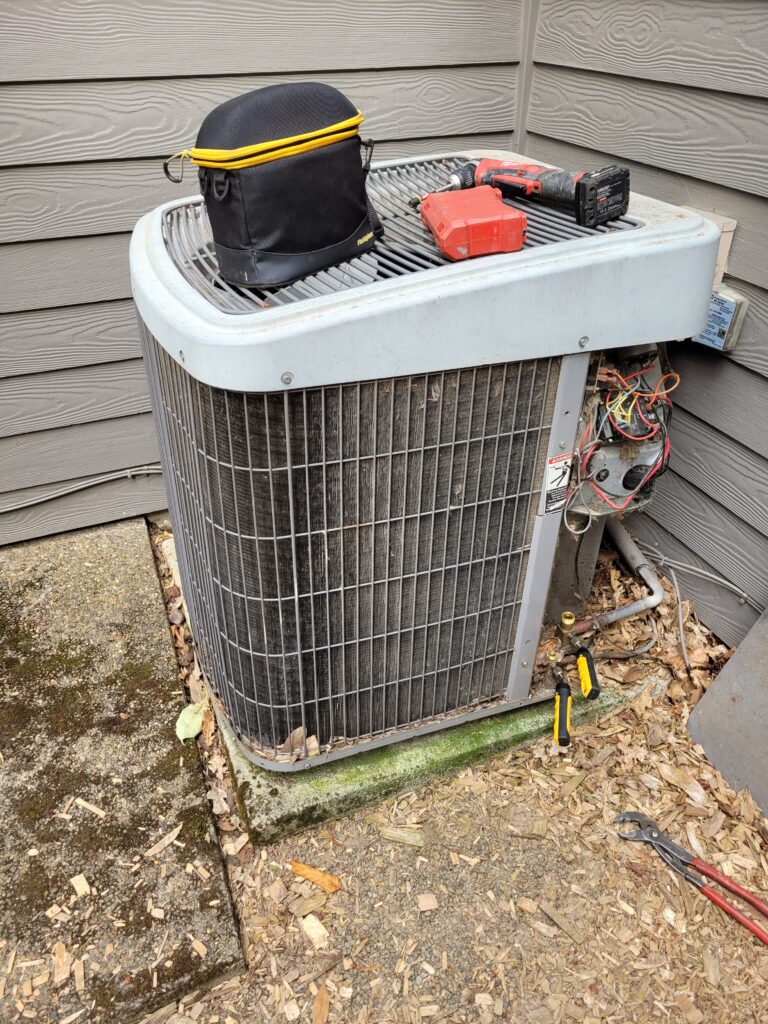 Outdoor AC condenser with electrical panel open, wiring exposed, and a drill and tool bag on top beside gray siding