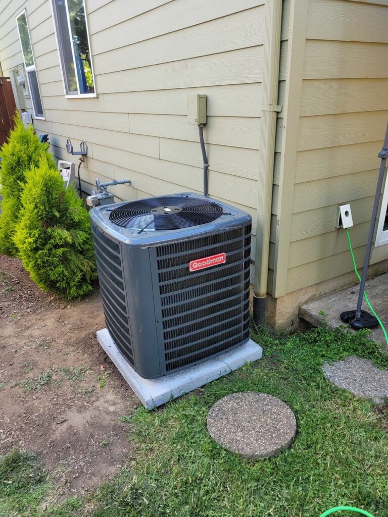 Gray Goodman air conditioner condenser on a concrete pad beside a tan-sided house, with disconnect box and shrubs nearby