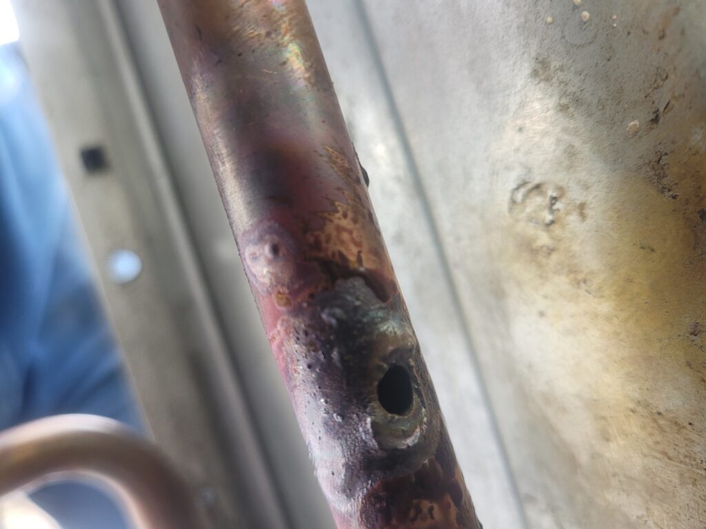 Close-up of a corroded copper refrigerant line with a hole and heat discoloration inside an HVAC unit cabinet