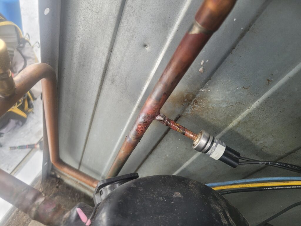 Copper refrigerant line with freshly brazed tee and pressure switch inside an outdoor condenser, compressor top in foreground