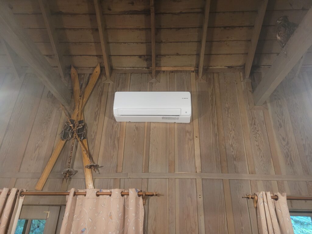 White ductless mini-split unit mounted high on a rustic wood-paneled cabin wall beside crossed vintage skis
