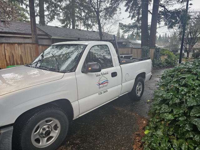 White pickup truck with a company logo on the door, parked on a wet driveway beside a hedge, wooden fence, and tall evergreens