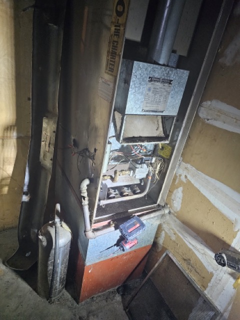 Older gas furnace with front panel removed, exposing burners and controls, in a cramped basement with a metal flue pipe