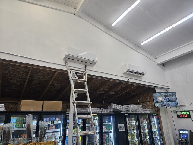 Two wall-mounted ductless mini-split units high on a white wall above store coolers, with an aluminum ladder beneath