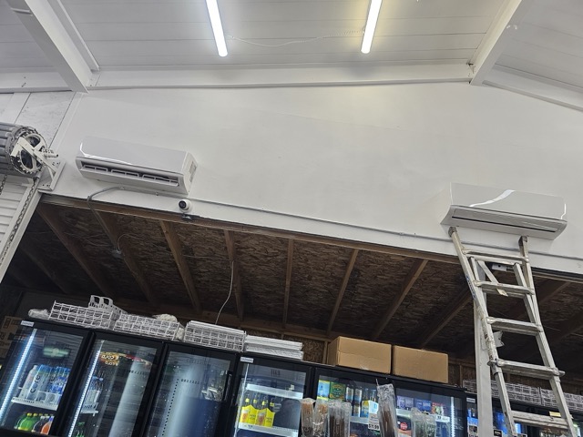 Two wall-mounted ductless mini-split units high on a white wall above store coolers, with an aluminum ladder below
