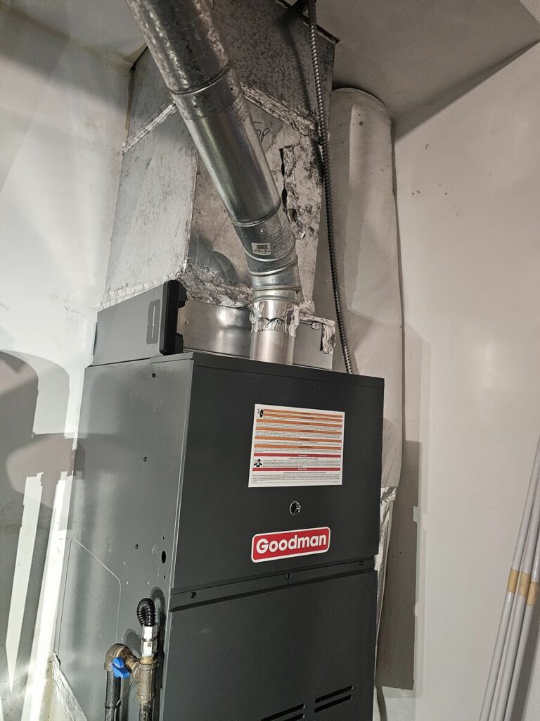 Gray Goodman gas furnace installed in a closet with galvanized flue pipe, sheet metal plenum, and gas line with blue shutoff valve