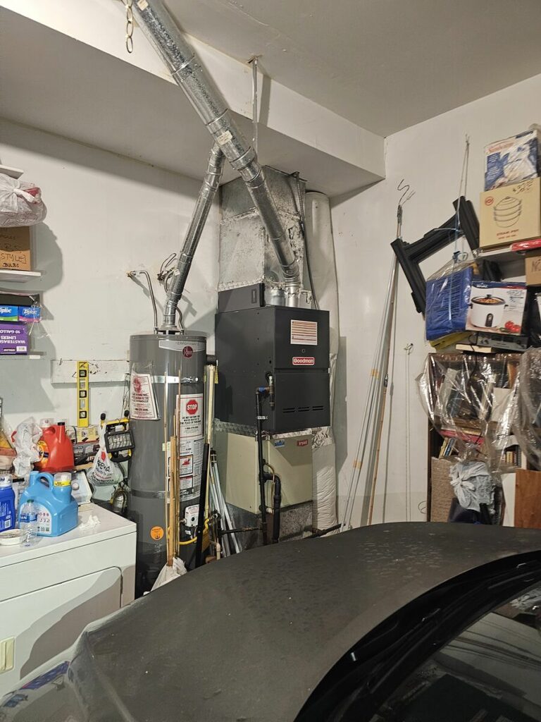 New black Goodman gas furnace with metal flue pipes beside a Rheem water heater in a residential garage