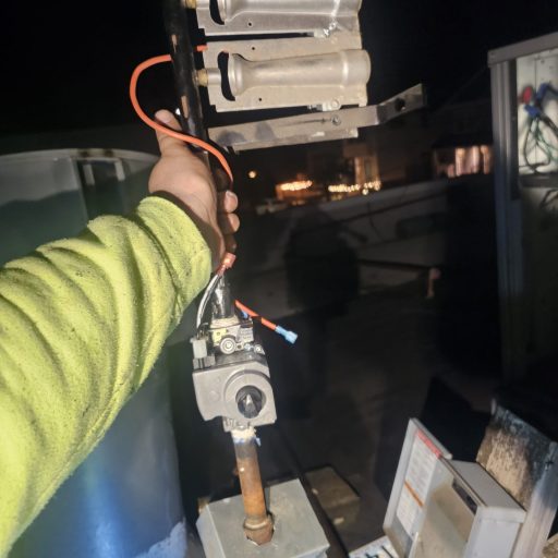 Technician holding a gas water heater burner assembly with multiple burner tubes, gas valve, and igniter wiring during a nighttime repair