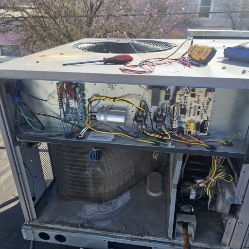 Open rooftop packaged HVAC unit undergoing service with multimeter and screwdriver on top, exposing control boards, capacitor, contactor, wiring, and condenser coil